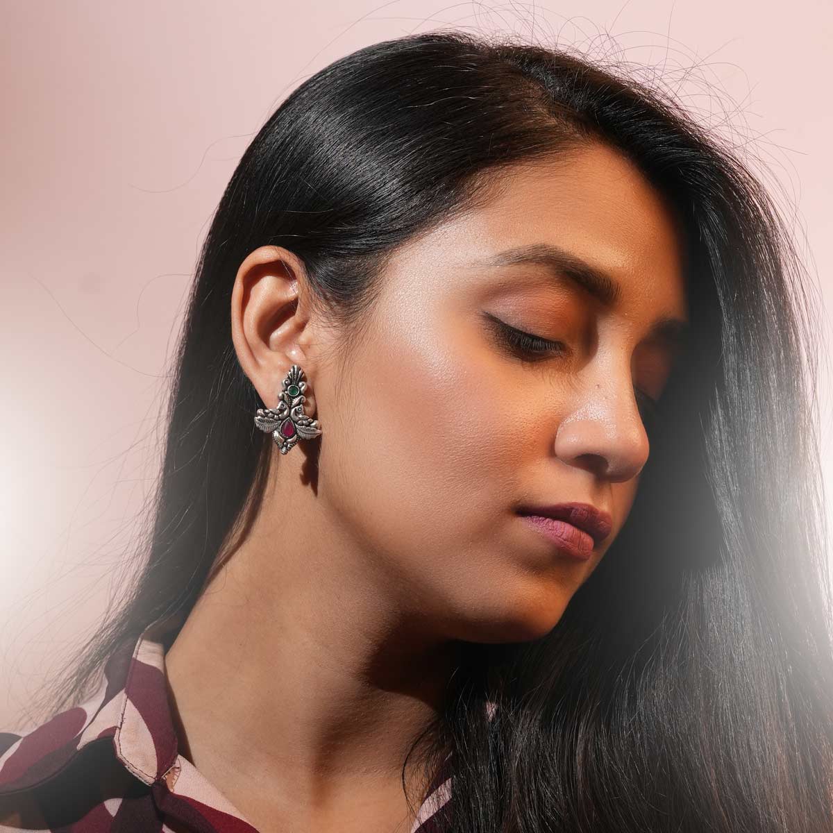 A pair of oxidized silver twin peacock earrings with intricate detailing and vibrant green and pink stones worn by a women.