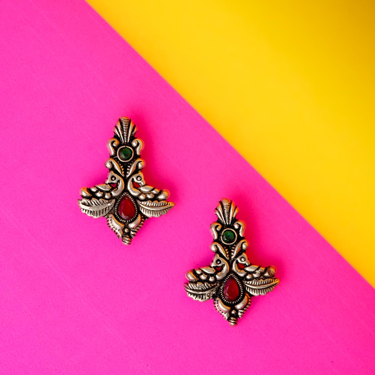 A pair of oxidized silver twin peacock earrings with intricate detailing and vibrant green and pink stones against a colourful background.