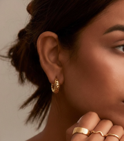 Close-up of a woman wearing gold hoop earrings and rings against a neutral background
