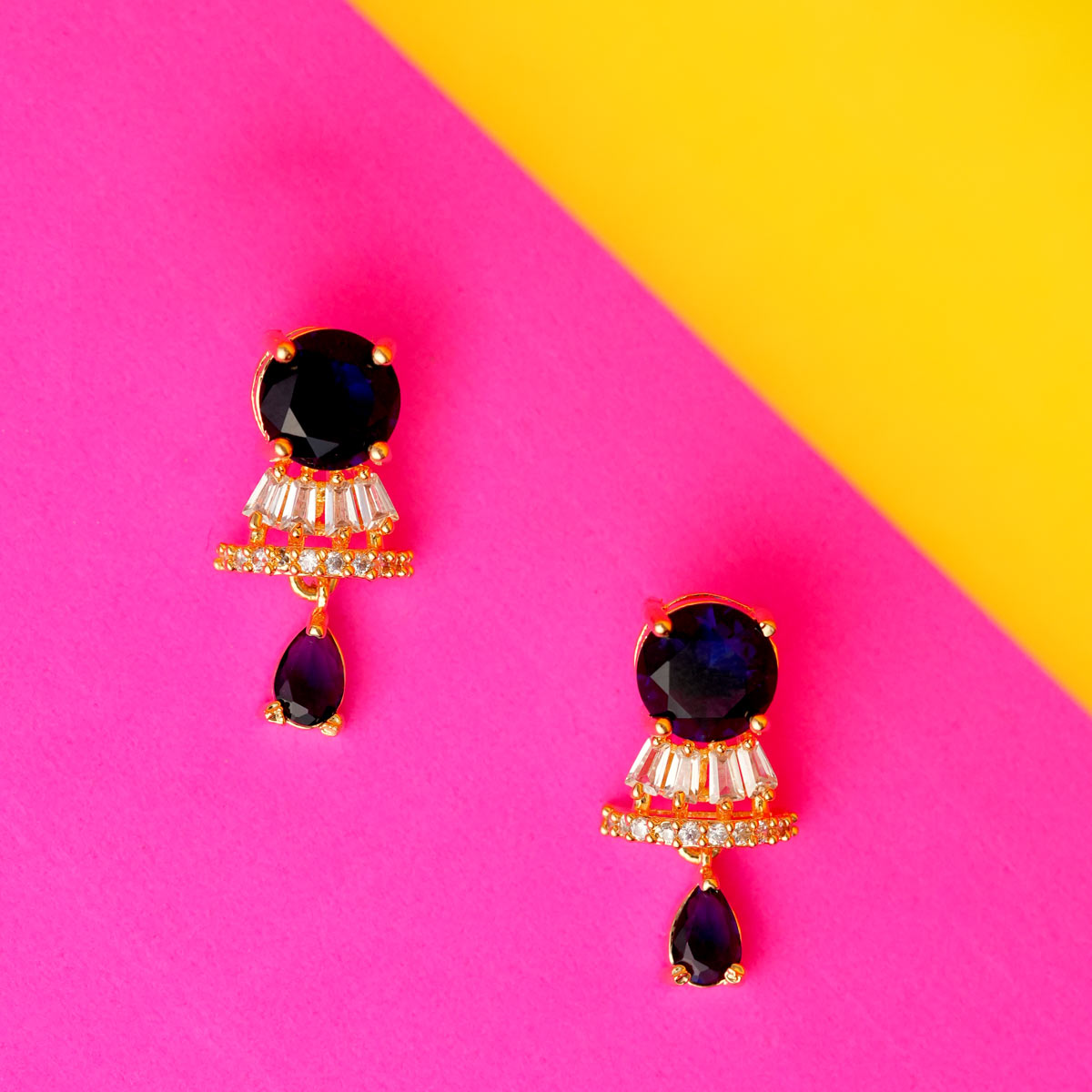 A pair of Midnight Sapphire Drop Earrings with round sapphire studs and cascading blue teardrop shapes with sparkling crystal accents on a colourful a background. 