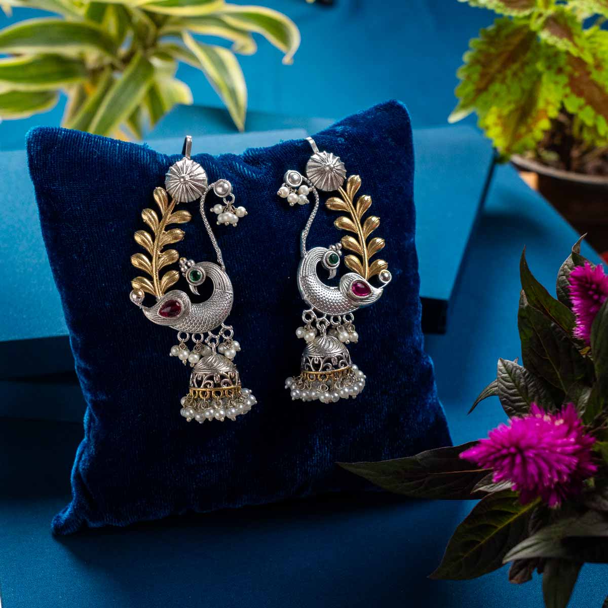 Peacock Decorative earrings on a blue pillow with a blue background and plants.