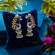 Peacock Decorative earrings on a blue pillow with a blue background and plants.